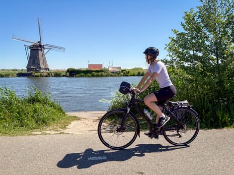 Female cyclist on path beside canal with traditional Dutch windmill in Kinderdijk under blue sky.