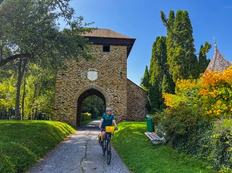Radfahrer mit gelben Taschen fährt durch gewölbtes Backsteintor am Murradweg. Grüne Wiese, Zypressen und Kirchturm im Hintergrund sichtbar.