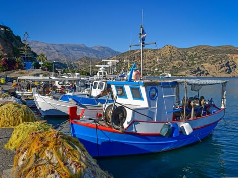 Blau-weiße Fischerboote im kretischen Hafen mit gelben Netzen am Kai. Berge und klarer blauer Himmel im Hintergrund sichtbar.