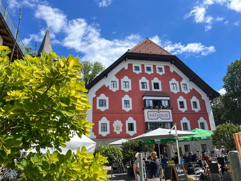 Red and white Rathaus building in Saalfelden with outdoor café seating under green umbrellas, framed by bright green foliage.
