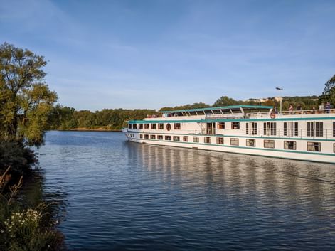 MS Switzerland Flusskreuzfahrtschiff mit weißem Rumpf und türkisfarbenen Akzenten auf ruhigem Fluss, umgeben von grünen Bäumen.