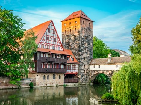 Medieval stone tower and half-timbered house beside river in Nuremberg, with stone bridge and weeping willows under blue sky.
