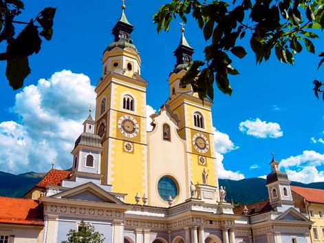 Brixen Cathedral with yellow and white twin towers, ornate clocks, and baroque facade under blue sky with white clouds and green foliage.
