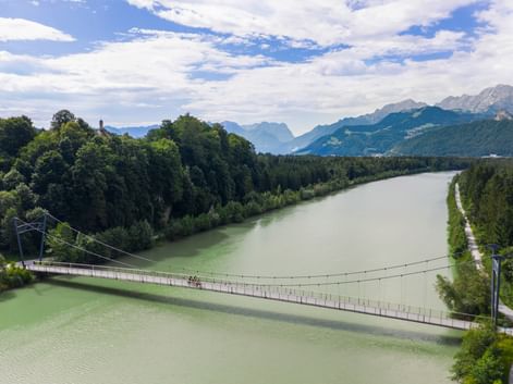 Suspension bridge crossing green river with cyclists, surrounded by forests and Alpine mountains under blue sky near Puch on Alpe-Adria route.