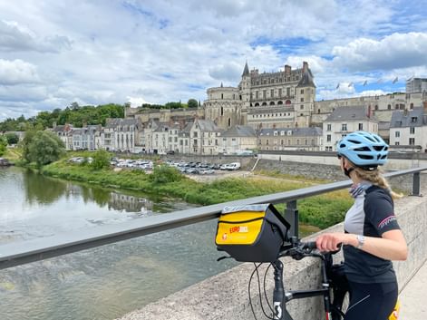 Radfahrerin mit blauem Helm auf Brücke mit Blick auf die Loire und Schloss Amboise. Das Renaissance-Schloss thront über historischen Gebäuden.