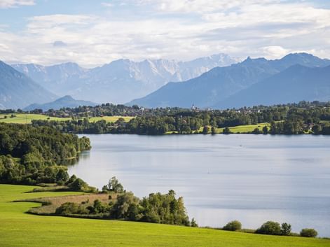 View of Riegsee lake surrounded by green meadows and forests, with snow-capped Alpine mountains in the background under a cloudy sky.