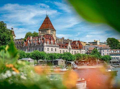 Schloss Château d'Ouchy mit rotem Ziegeldach und Turm in Lausanne am Genfersee, eingerahmt von grünem Laub und bunten Blumen im Vordergrund.
