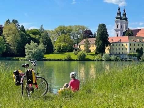 Radfahrer in rotem Shirt und Helm sitzt am See mit Fahrrad. Gelbe Klostergebäude und Kirche mit Doppeltürmen am gegenüberliegenden Ufer.