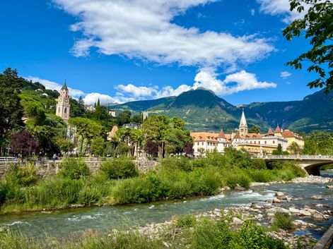 Die Passer fließt durch Meran mit historischen Gebäuden, Kirchtürmen und Bergen in Südtirol unter blauem Himmel mit weißen Wolken.