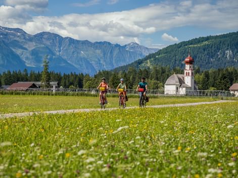 Drei Radfahrer auf einem Weg durch eine Blumenwiese in Leutasch, mit weißer Kirche mit roter Zwiebelkuppel und Alpenbergen im Hintergrund.