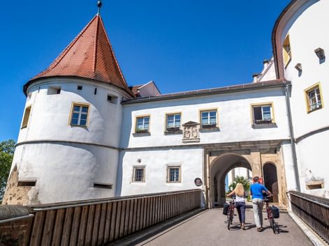 Zwei Radfahrer mit Rädern an der Eingangsbrücke von Schloss Wörth, einem weißen Gebäude mit rotem Dach und Rundturm unter blauem Himmel.