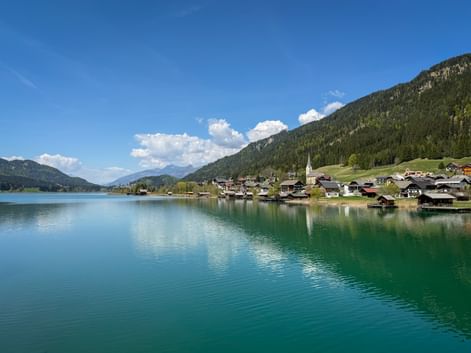 Turquoise Weissensee lake with Techendorf village on the shore, featuring a church spire and traditional houses against forested mountains.
