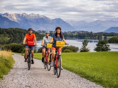 Three cyclists with helmets riding along Riegsee lake in Bavaria. Mountains and green meadows visible in background under cloudy sky.