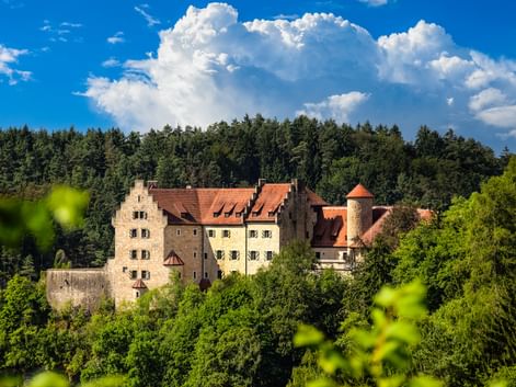 Burg Rabenstein mit roten Ziegeldächern und rundem Turm, umgeben von grünem Wald unter dramatischen weißen Wolken und blauem Himmel in Österreich.