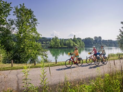 Three cyclists riding along a paved path beside a calm green river with trees and forest in the background under a blue sky.