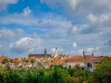 Panoramic view of Rothenburg ob der Tauber with medieval buildings, orange-tiled roofs, and church towers above green trees under a cloudy sky.