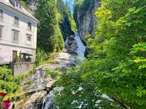 Waterfall cascading through rocky gorge in Bad Gastein, framed by green trees and a white building on the left.