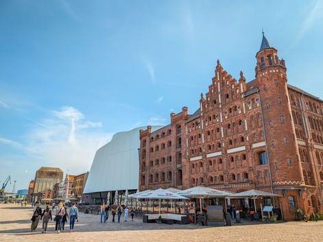 Red brick Türmchenspeicher building with tower in Stralsund, adjacent to modern white structure. People gather on the plaza under blue sky.