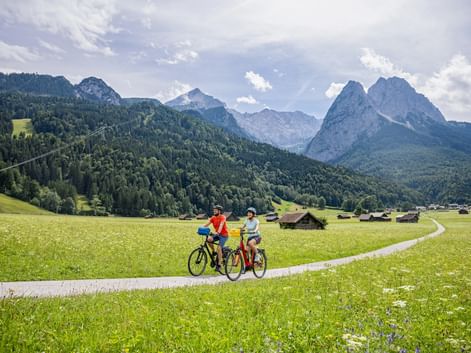 Two cyclists on a path through green meadows near Garmisch with traditional wooden houses and dramatic Alpine peaks in the background.