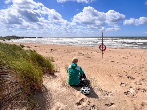 Radfahrer in türkisfarbenem Shirt sitzt am Sandstrand mit Helm neben sich und blickt aufs Meer. Dünengras links, Rettungsring am Strand.