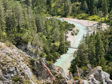 Aerial view of turquoise Isar River winding through dense forest with rocky banks and a small footbridge crossing the water.