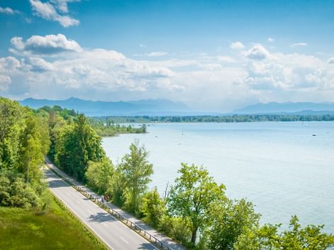 Scenic cycling path along Lake Starnberg with lush green trees, calm blue water, and mountain ranges in the distance under a cloudy sky.
