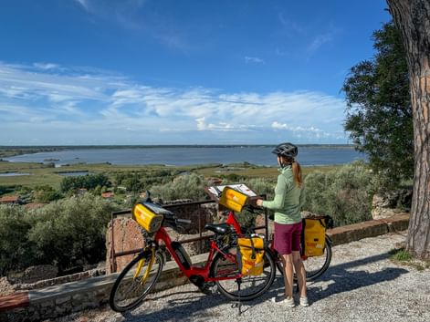 Radfahrerin mit rotem Tourenrad und gelben Packtaschen mit Blick auf den Lago di Massaciuccoli in der Toskana unter blauem Himmel.