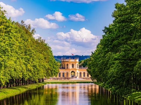 Yellow Orangerie palace in Kassel framed by symmetrical rows of green trees along a reflective canal under a blue sky with white clouds.