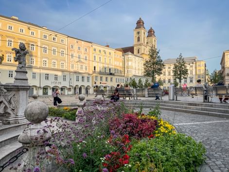 Linzer Hauptplatz mit bunten Blumenbeeten im Vordergrund, gelben historischen Gebäuden und Kirche mit zwei Türmen unter blauem Himmel.
