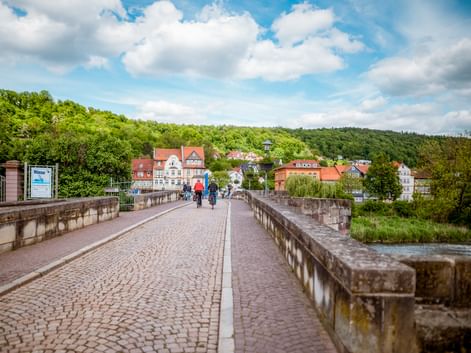 Two cyclists riding across historic cobblestone Werra bridge in Hann. Münden with colorful half-timbered houses and green hills in background.