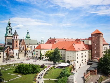 Aerial view of Wawel Castle complex in Krakow with red-roofed buildings, cathedral with green copper towers, and round brick tower.