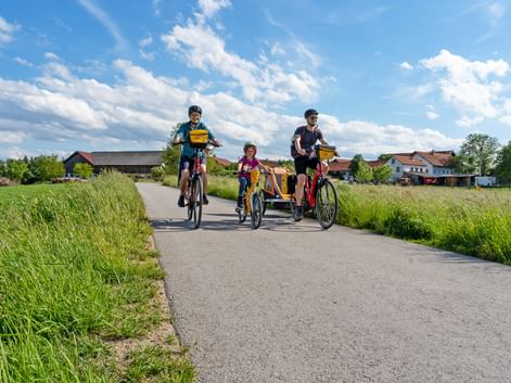 Family of four cycling on paved path through green countryside. Two adults and two children with trailer bike under blue sky with clouds.