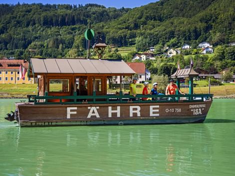 Fähre mit Radfahrern überquert die grüne Donau bei Engelhartszell. Bewaldete Hügel und Dorfgebäude im Hintergrund sichtbar.