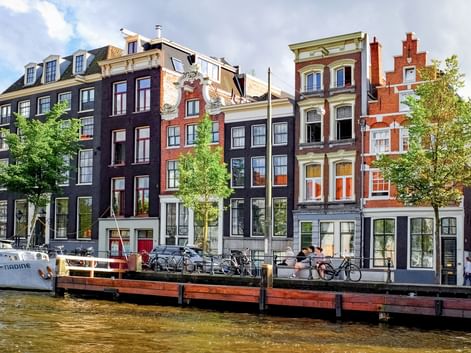 Row of traditional Amsterdam canal houses in various colors along a waterway. Bicycles are parked on a wooden dock in front of the buildings.
