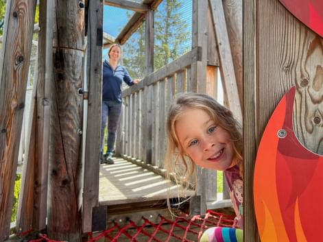Smiling girl peeks through wooden playground structure while woman stands on elevated walkway in background. Sunny day with blue sky and trees.