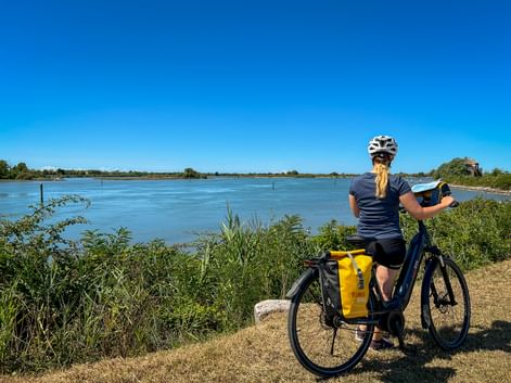 Radfahrerin mit Helm und gelben Packtaschen am Fahrrad blickt auf breiten Fluss unter blauem Himmel auf der Route Venedig-Mantua.