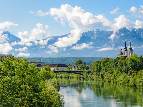 Blick auf die Drau in Villach mit einer Kirche mit zwei Türmen, grünen Bäumen und schneebedeckten Bergen unter blauem Himmel.