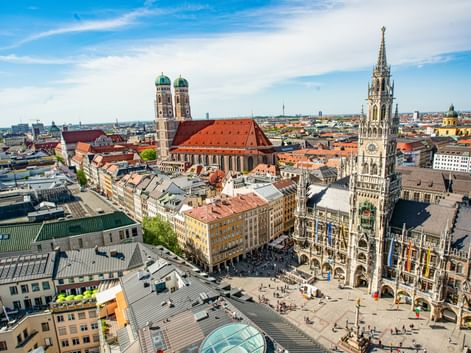Aerial view of Munich's historic center showing Marienplatz with the New Town Hall tower and Frauenkirche's twin domes under blue sky.