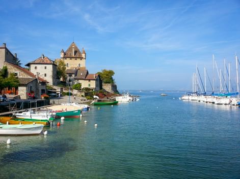 Malerischer Hafen von Yvoire am Genfersee mit bunten Booten, mittelalterlichen Schlosstürmen und Segelbooten unter blauem Himmel.