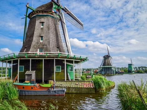 Traditionelle niederländische Windmühle mit grünem Rand neben einem Kanal in Kinderdijk. Ein kleines Boot liegt am Fuß vertäut, weitere Windmühlen sind in der Ferne sichtbar.