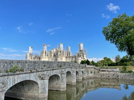 Stone bridge with multiple arches spanning calm water, leading to Château de Chambord with its distinctive towers under blue sky.