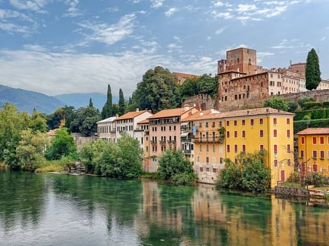Farbenfrohe historische Gebäude von Bassano del Grappa spiegeln sich im Fluss Brenta, mit einer mittelalterlichen Burg auf dem Hügel.