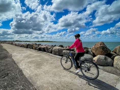 Radfahrerin in rosa Jacke mit Fahrrad auf gepflastertem Küstenweg neben steinerner Uferbefestigung. Blaues Meer und dramatischer Wolkenhimmel im Hintergrund.