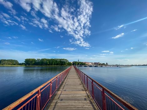Long wooden pier with red railings extending across calm blue water near Cammin, with green shoreline and town buildings visible in the distance.
