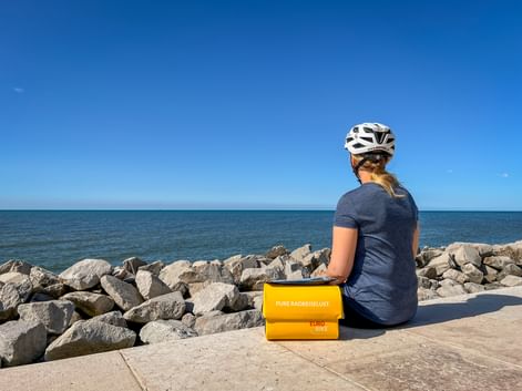 Radfahrerin mit weißem Helm sitzt auf der Promenade in Lido di Venezia mit Blick aufs Meer. Gelbe Eurobike-Taschen neben ihr, Steinmole sichtbar.