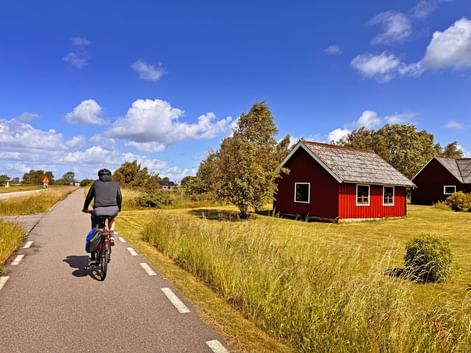 Radfahrer auf asphaltiertem Radweg entlang Sydkustleden in Schweden, vorbei an traditionellem roten Holzhaus mit weißen Rahmen unter blauem Himmel.