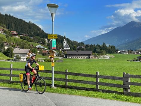 Radfahrer am Tauernradweg bei Krimml mit grünen Wiesen, Alpendorf mit Kirche, Holzzaun und Bergen unter blauem Himmel.