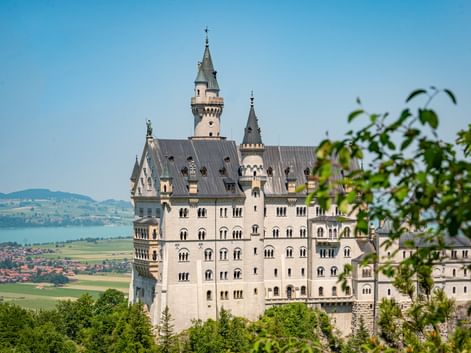 Schloss Neuschwanstein mit weißen Mauern und grauen Türmen umgeben von grünen Bäumen. See und Dorf im Hintergrund unter blauem Himmel.
