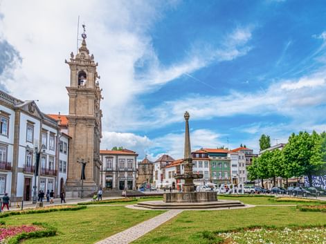 Largo Carlos Amarante in Braga with bell tower, fountain, green lawns, and colorful buildings under blue sky.