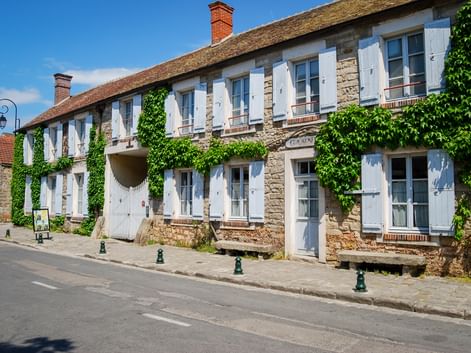 Two-story stone building in Barbizon with white shutters and ivy-covered walls. The facade features multiple windows and a central entrance.